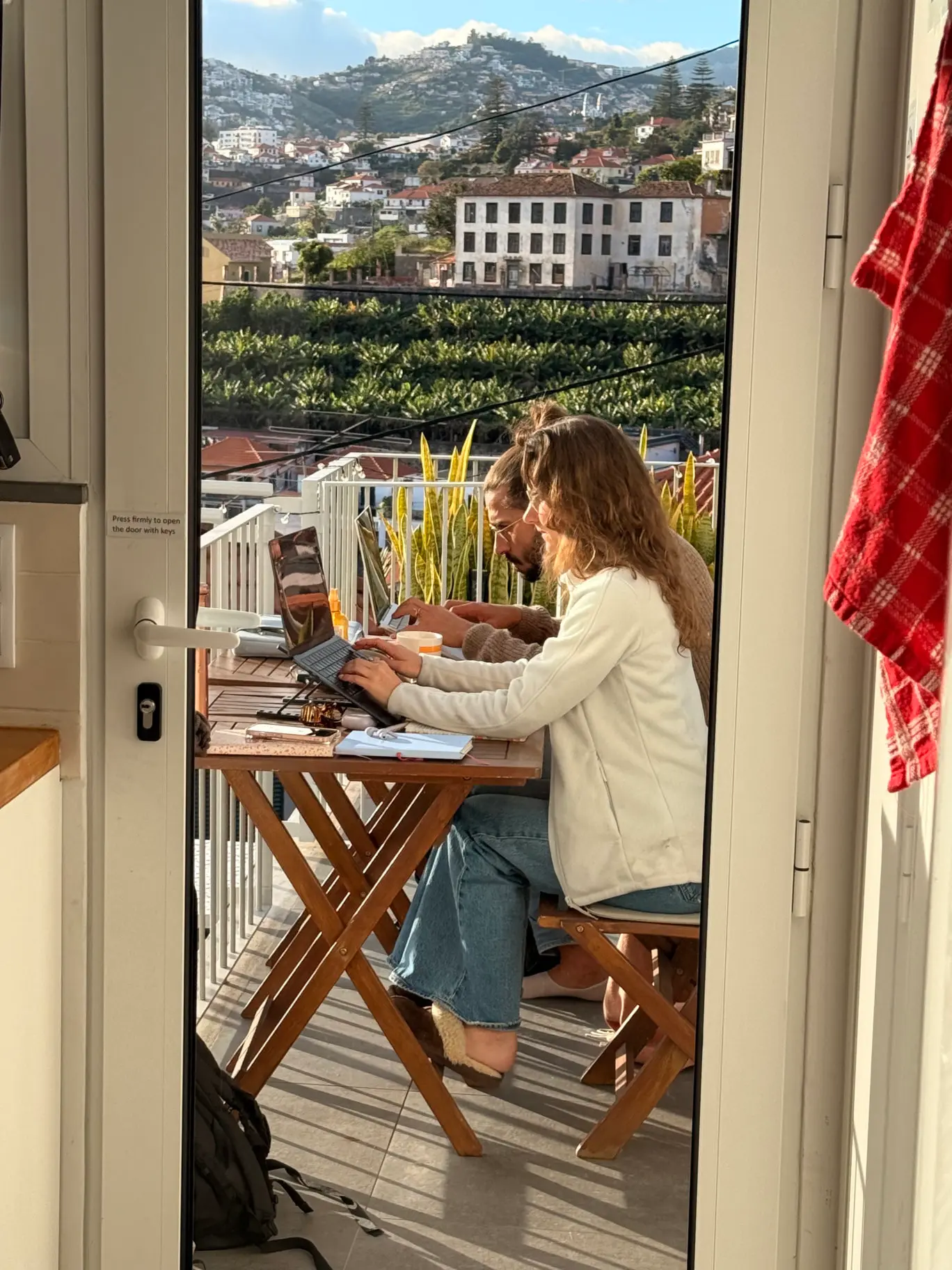 Two residents working on laptops at a table on the balcony, seen through the kitchen door, with a view over Funchal